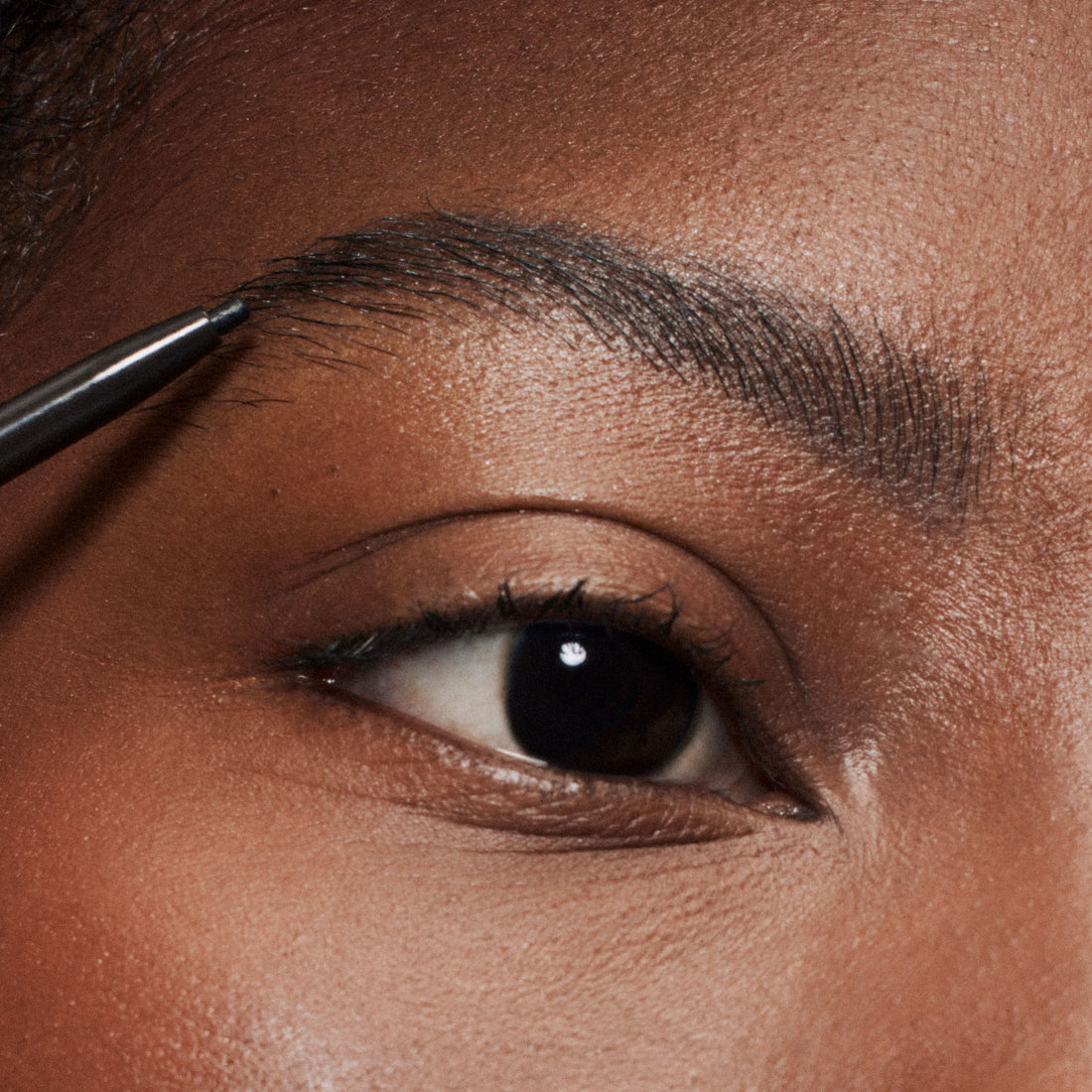 A close up image of a model applying ILIA's Brow Pencil in soft black  to their brows. 