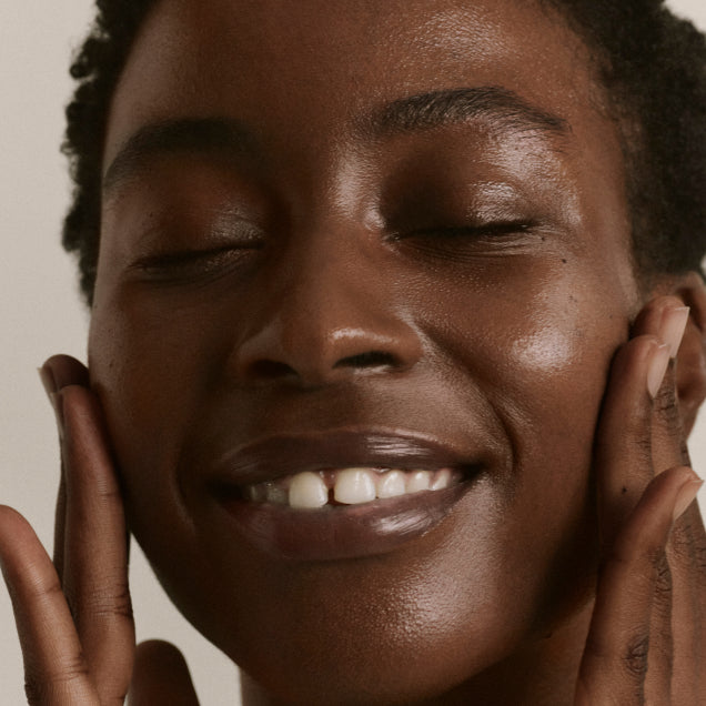 A model applying ILIA's Base Face Milk to their face with both hands, smiling with quenched skin.