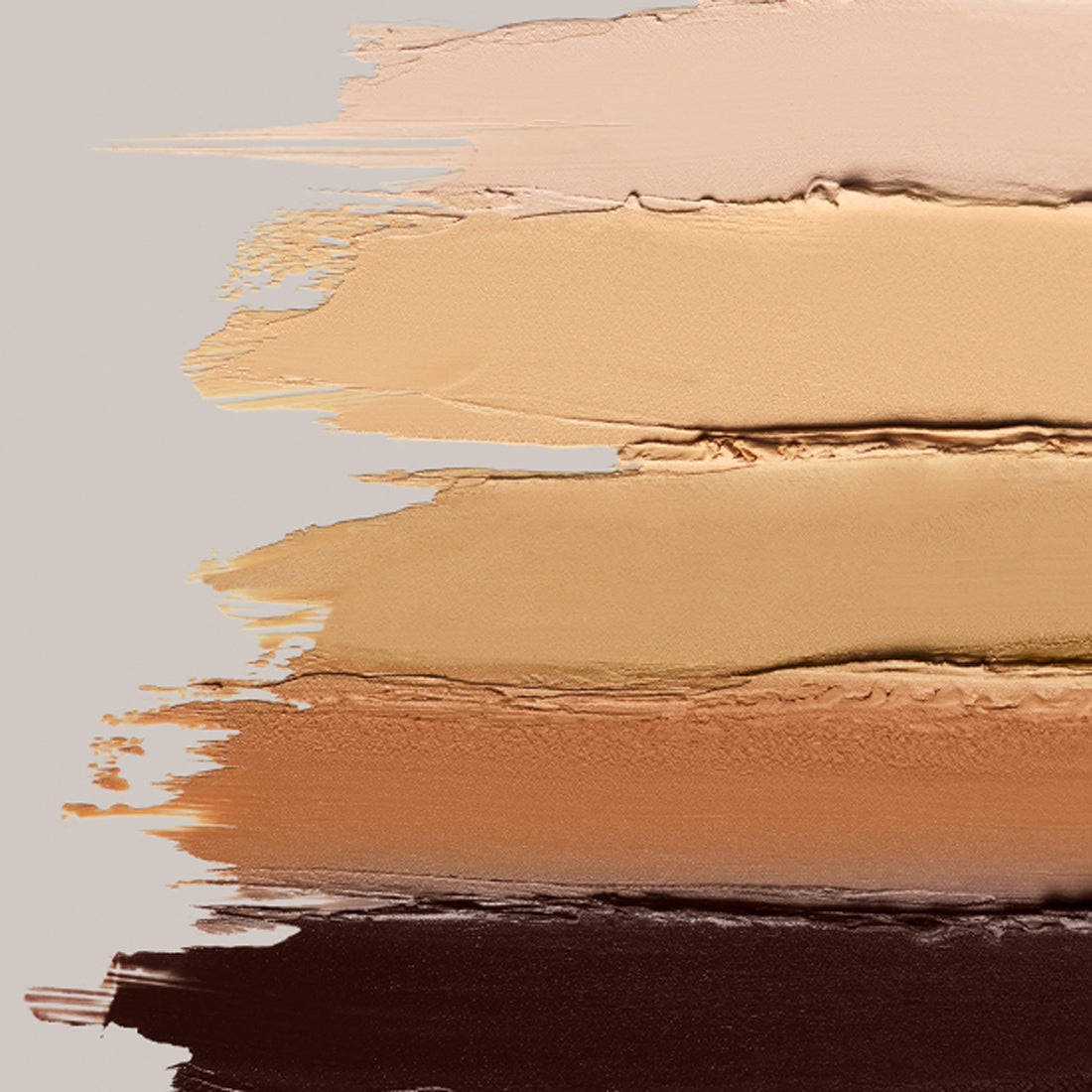 An array of ILIA's Skin Rewind Complexion Stick creamy swatches lined up against one another on a grey background.