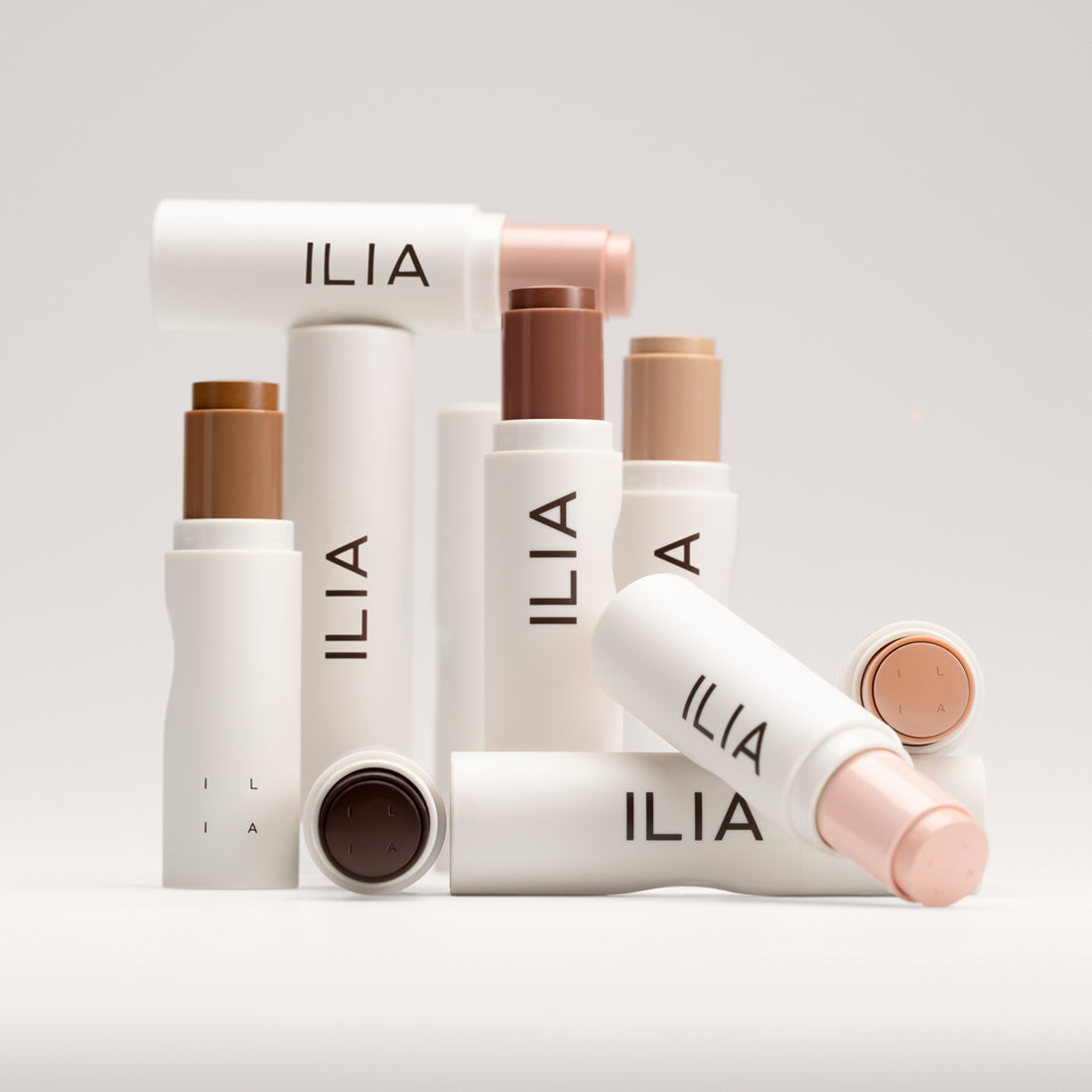 A variety of ILIA's Skin Rewind Complexion Stick shades styled on a grey-white background.