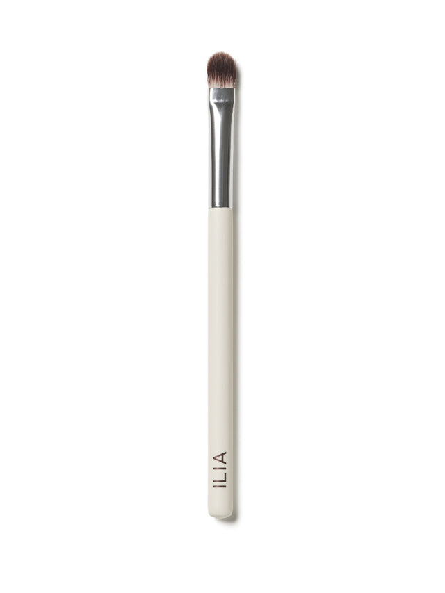 A close up of ILIA Shadow Brush that has a flat, tapered head and a white handle.