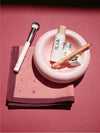 ILIA makeup products including a brush, complexion stick, lip pencil, sharpener, and lip + cheek stick on a pink plate and scarf.