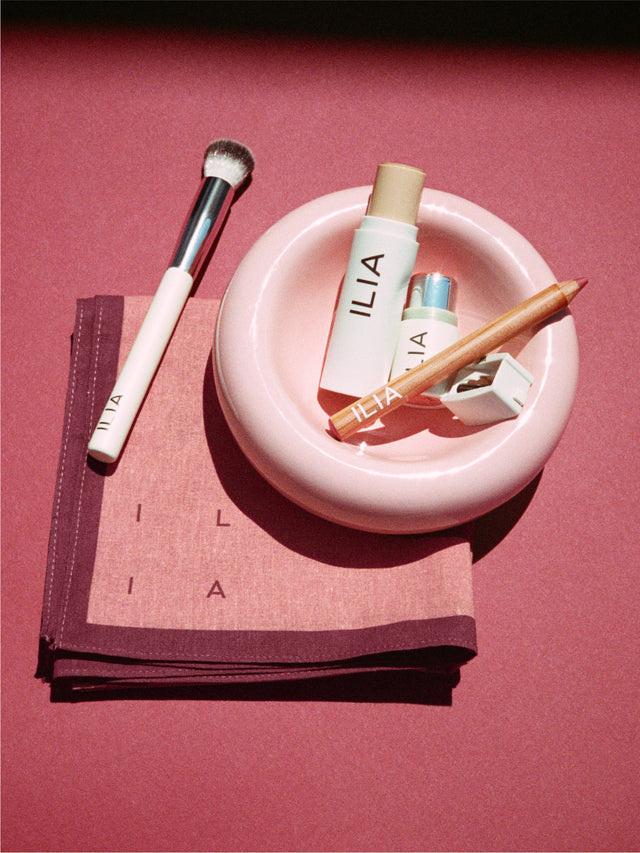 ILIA makeup products including a brush, complexion stick, lip pencil, sharpener, and lip + cheek stick on a pink plate and scarf.